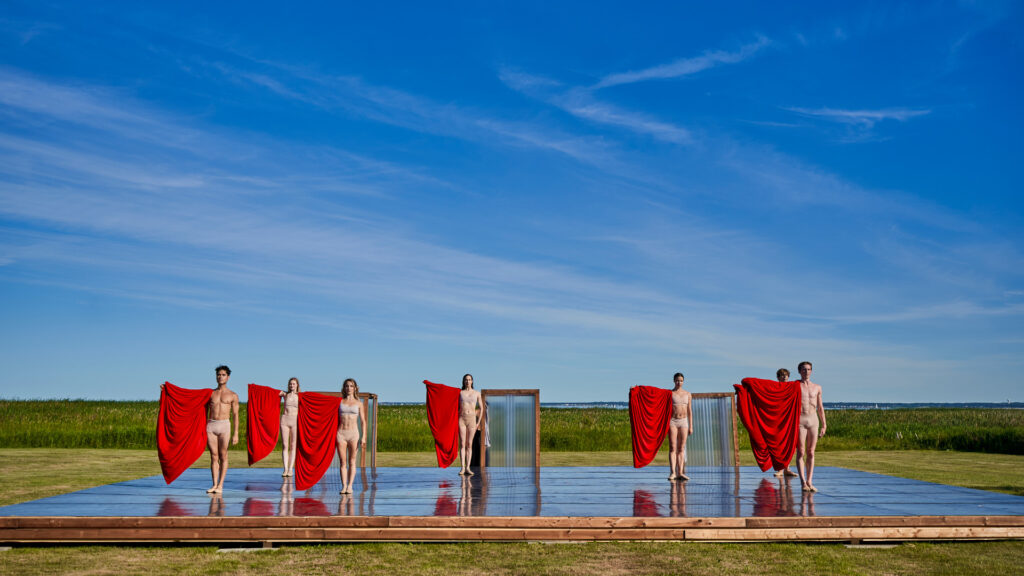 Seven dancers stand still on a wooden stage set up by the seashore.