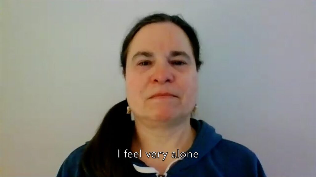 Still from a video showing the face of a woman in front of a white wall. The caption reads "I feel very alone".