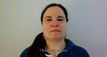 Still from a video showing the face of a woman in front of a white wall. The caption reads "I feel very alone".