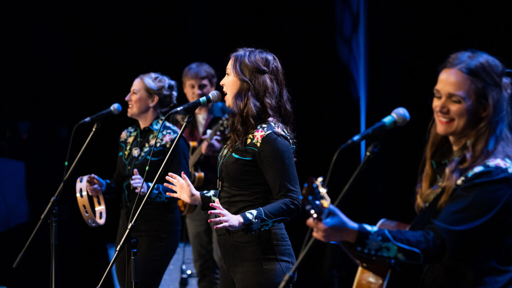 Three women sing and play joyously, along with a guitarist at the back. The side angle from the photo could lead to believe that the performers were close to one another, but physical distancing is implemented.
