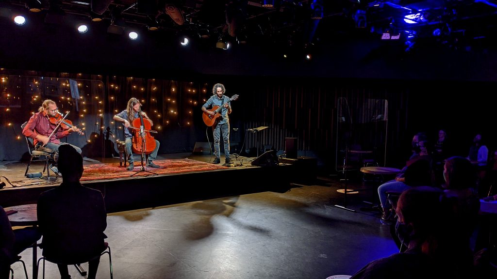 A violin, guitar and cello trio playing in a small indoor venue, with audience members wearing masks.