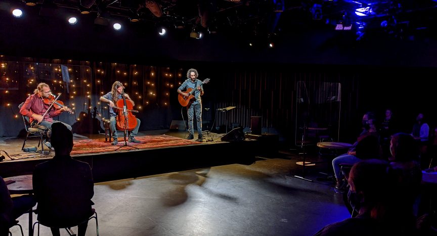 A violin, guitar and cello trio playing in a small indoor venue, with audience members wearing masks.