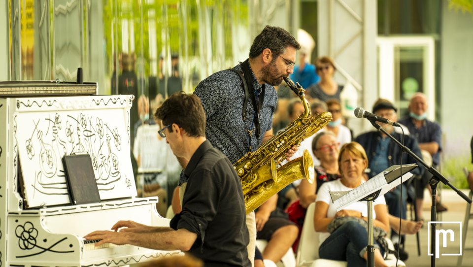 Vue de côté d'un pianiste et d'un saxophoniste s'exécutant devant un public visible en arrière-plan ainsi que dans les fenêtres qui offrent un effet miroir.