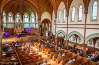 Two musicians performing before a socially distanced audience in a wooden church building.