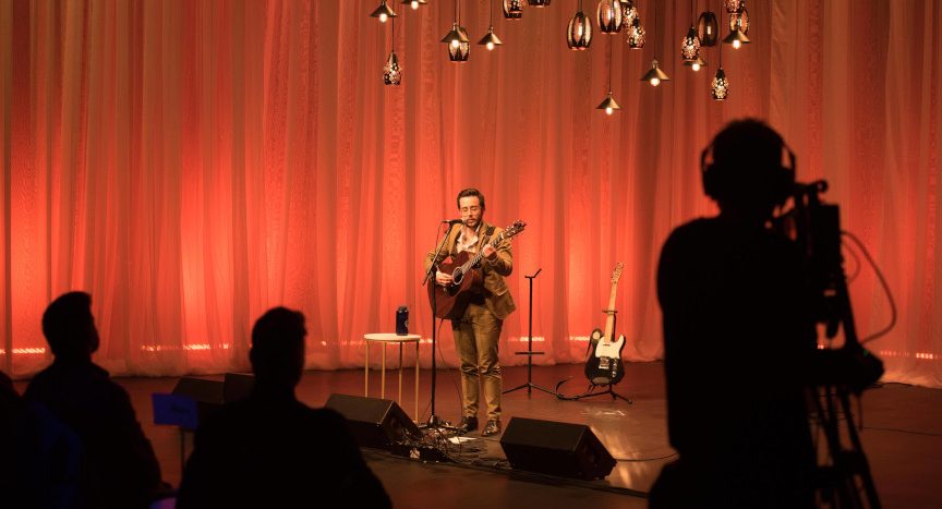 Photo d'un musicien seul sur scène avec sa guitare. À l'avant-plan, on aperçoit les silhouettes du public et d'un caméraman réalisant une captation du spectacle.