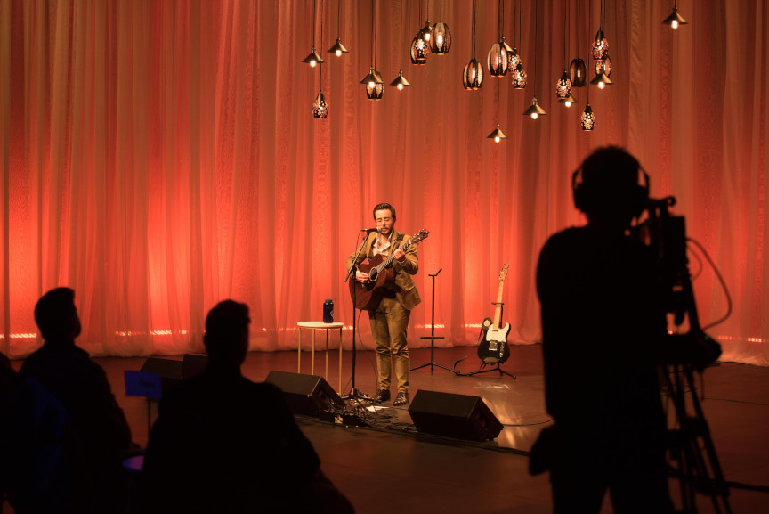 A singer performing with a guitar. On the foreground, dark silhouettes of audience members and of a technician recording the performance.