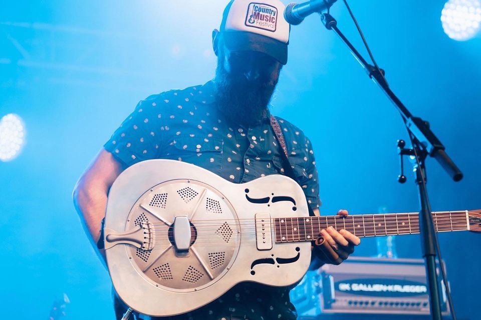 Un musicien se tient en arrière d'un microphone. Il y a des lumières bleues en arrière-scène. Le musicien tient une guitare blanche. Il porte un chapeau bleu et blanc et regarde vers le bas.