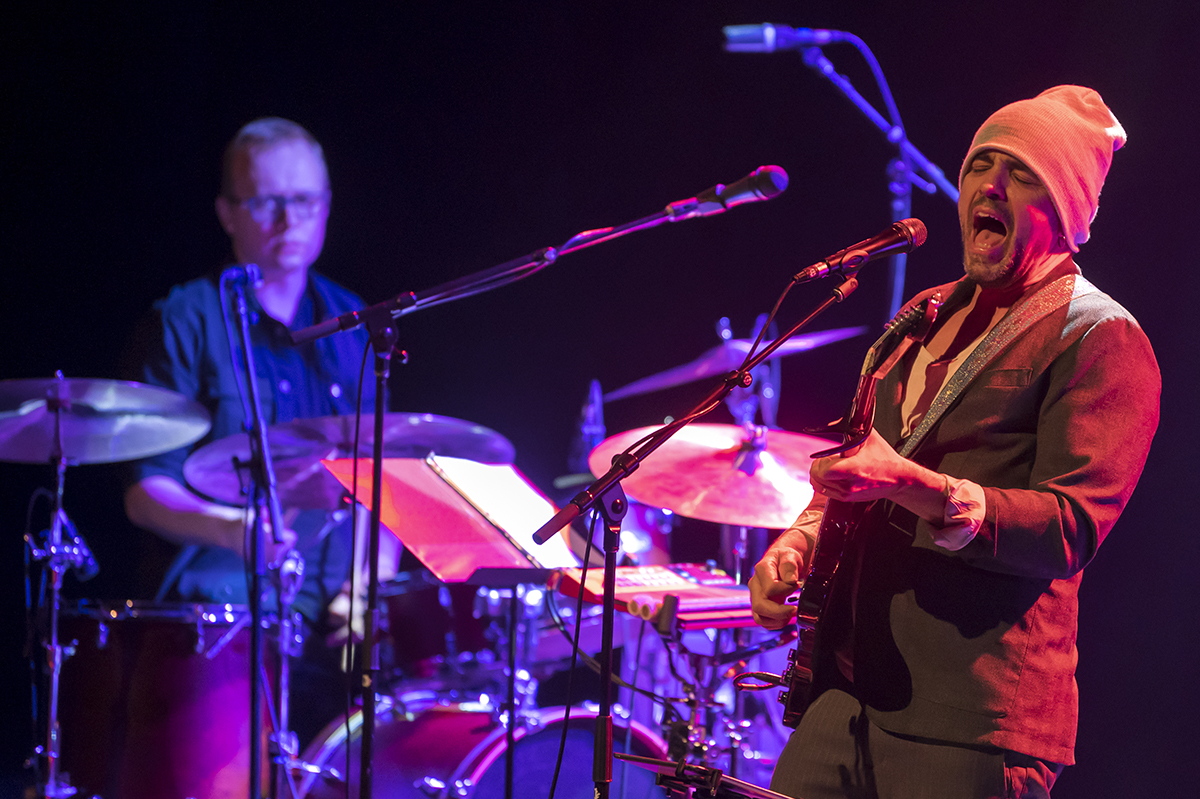 Musician singing and playing guitar, with a drummer in the background.