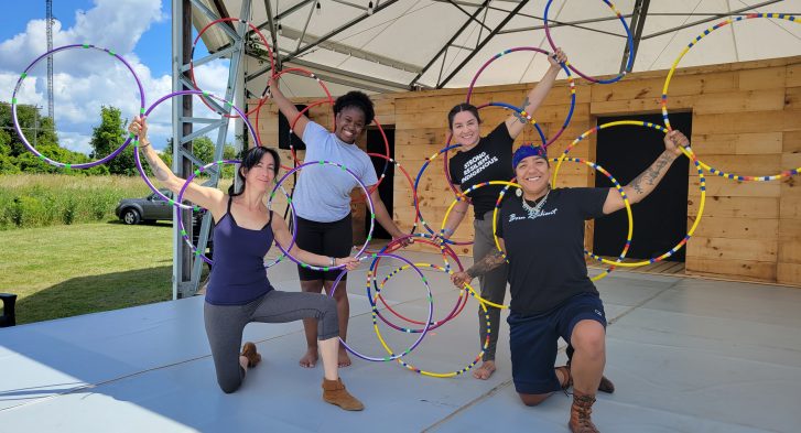 Four people form a semicircle on stage, smiling and holding a series of hoops in their outstretched hands.