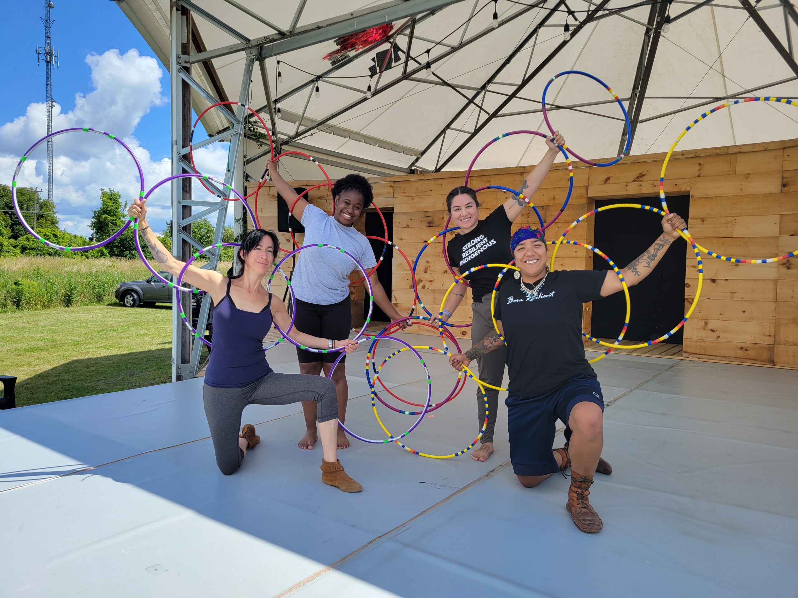 Four people form a semicircle on stage, smiling and holding a series of hoops in their outstretched hands.