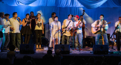 A group of musicians and singers lined up on a stage, performing, with blue curtains as a backdrop.
