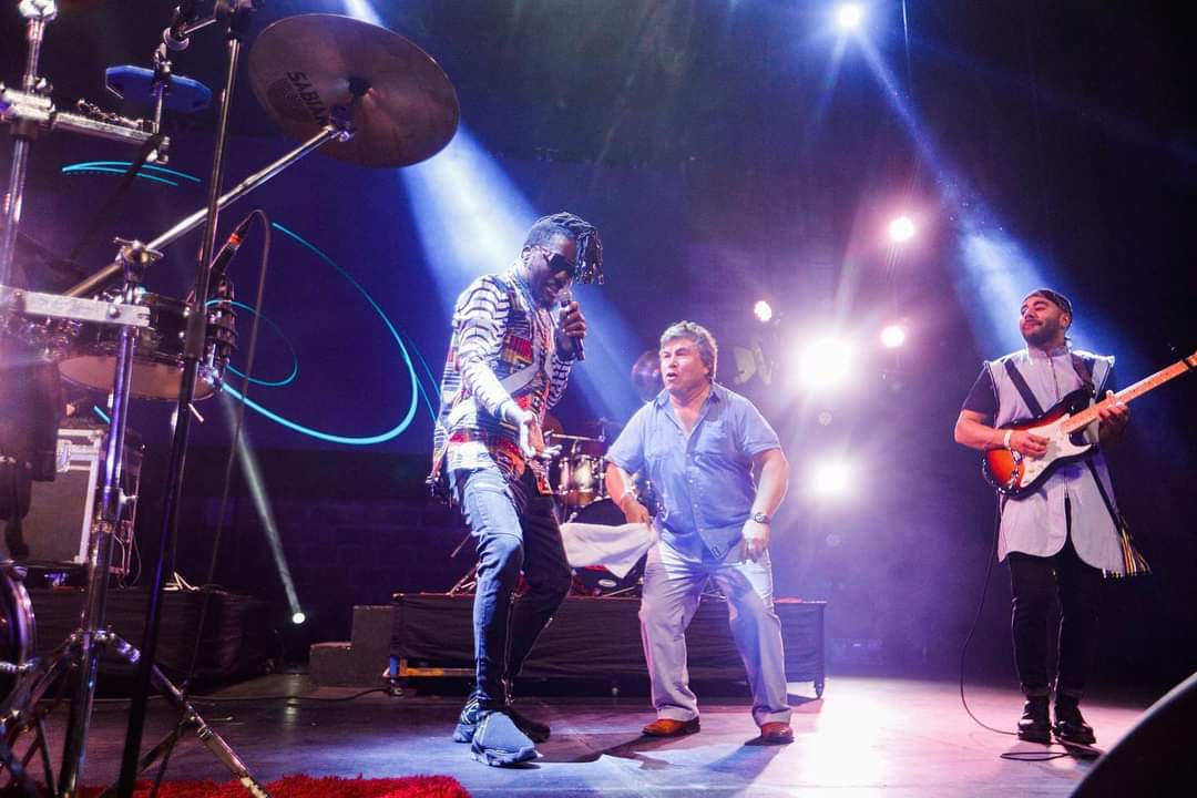 KIZABA and his band playing and singing with Alfredo on stage. The stage is covered in blue, purple and pink stage lights shining over the stage.