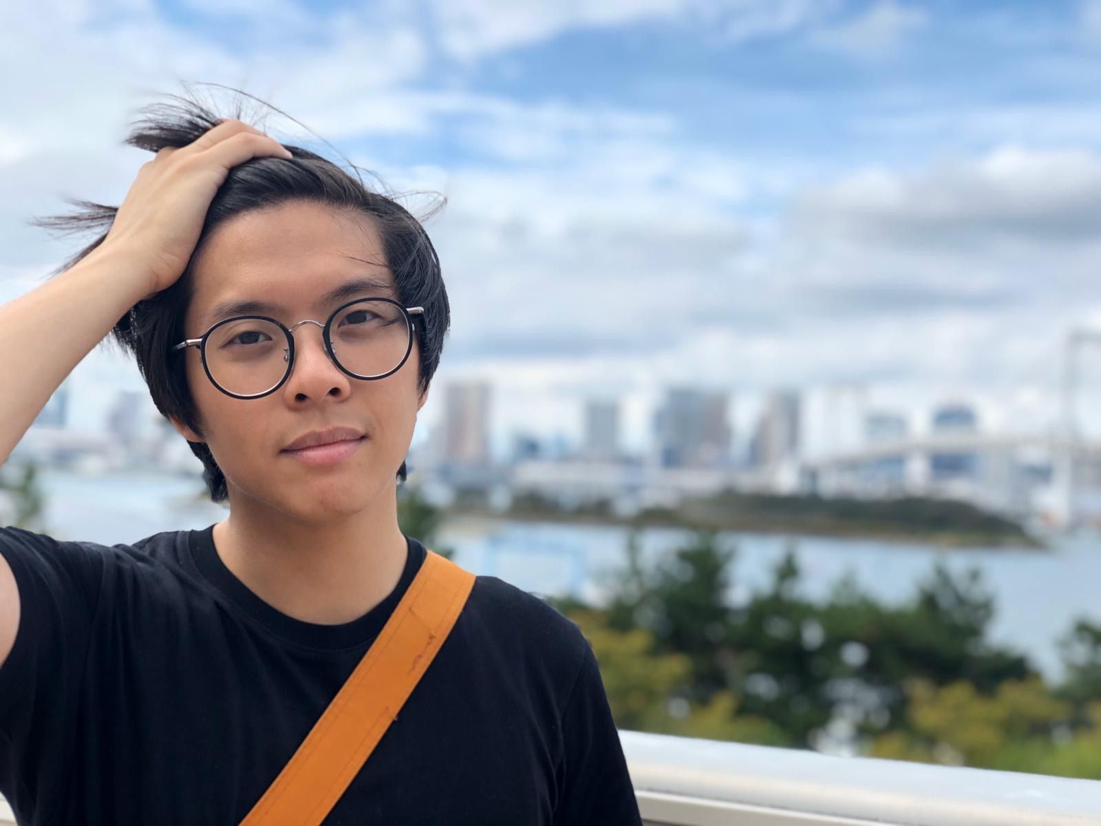 Une photo de Milton passant sa main droite dans ses cheveux. En arrière-plan, on peut voir un plan d'eau, l'horizon d'une ville et des arbres verts.