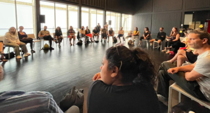 Un groupe de délégué(e)s canadien(ne)s qui rencontrent des artistes locaux. Ils sont assis en cercle, dans une pièce éclairée par des lumières vives, avec des murs et un plafond noirs, un sol sombre et un mur de fenêtres.