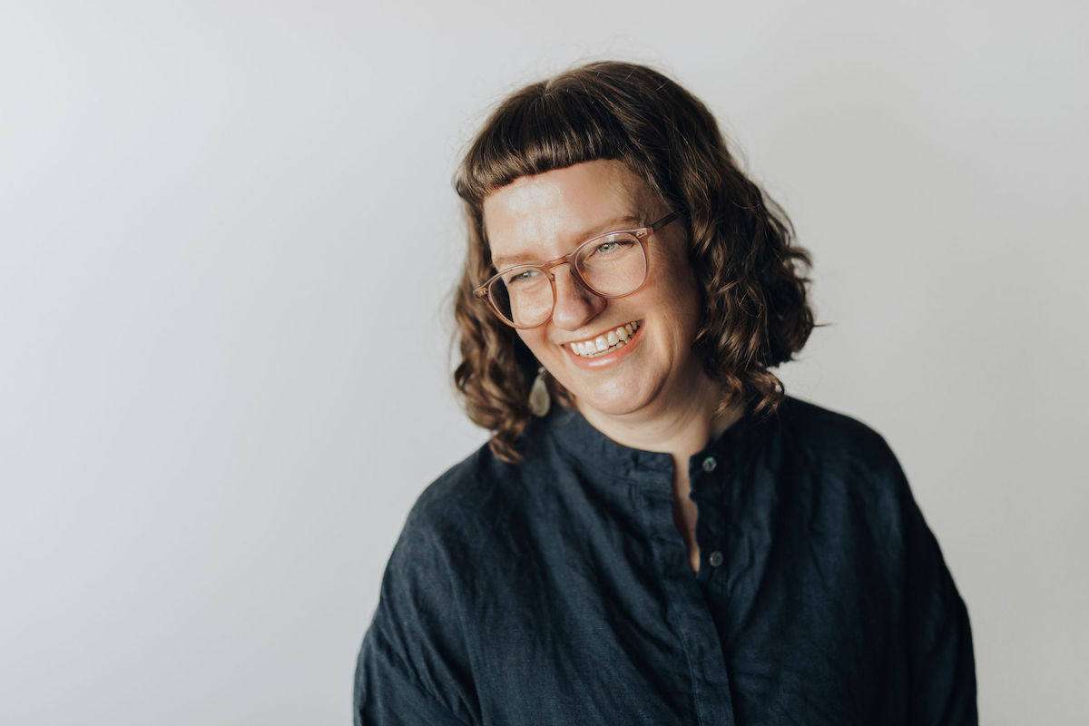 Portrait de Kristina Lemieux. Elle a des cheveux bruns bouclés à longueur d'épaule, des lunettes et porte une chemise vert foncé avec des boutons, devant un fond gris.