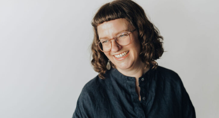 Headshot of Kristina Lemieux. She has shoulder-length curly brown hair, glasses, and is wearing a dark green shirt with buttons, in front of a grey background.