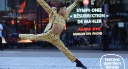 Un danseur habillé d'un costume à carreaux bondit dans la rue, devant une fenêtre réfléchissante.