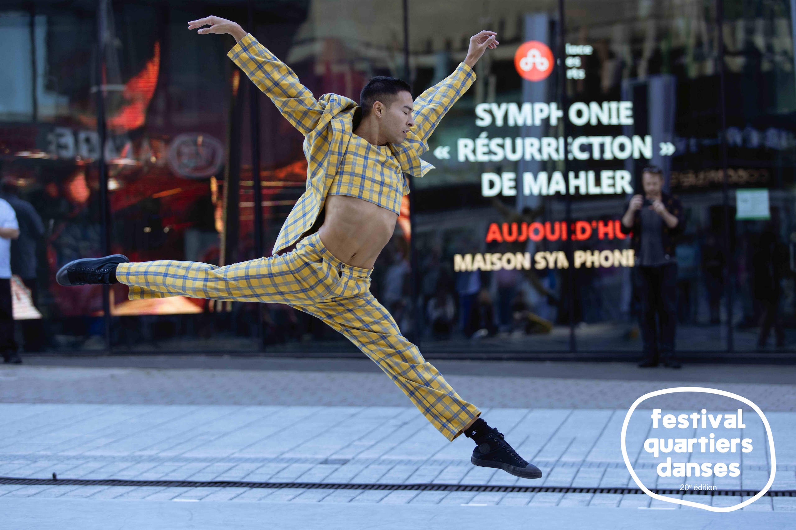 Un danseur habillé d'un costume à carreaux bondit dans la rue, devant une fenêtre réfléchissante.