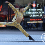 A dancer dressed in a plaid suit leaps on the street, in front of a reflective window.