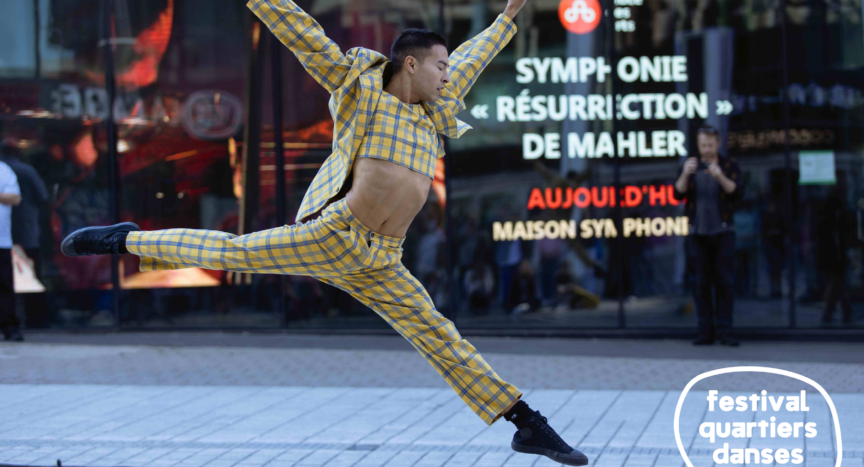 A dancer dressed in a plaid suit leaps on the street, in front of a reflective window.
