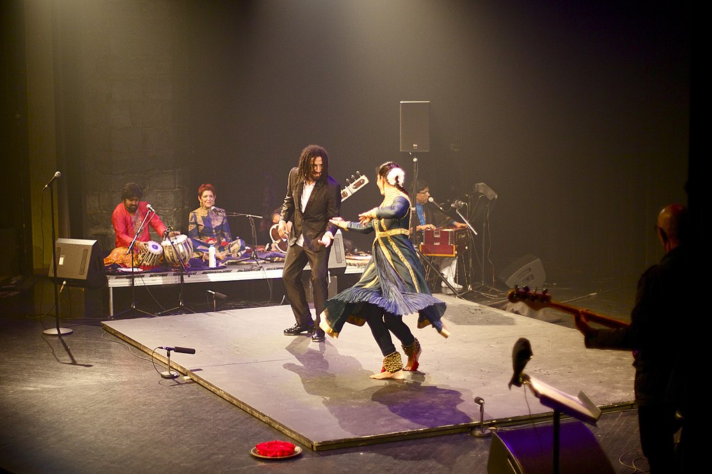 Two dancers are performing on a stage. The stage is surrounded by musicians.