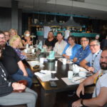 Dans un restaurant bondé, un groupe de délégués est assis autour d'une grande table en souriant pour une photo. Le(la) photographe prend la photo du bout de la table.