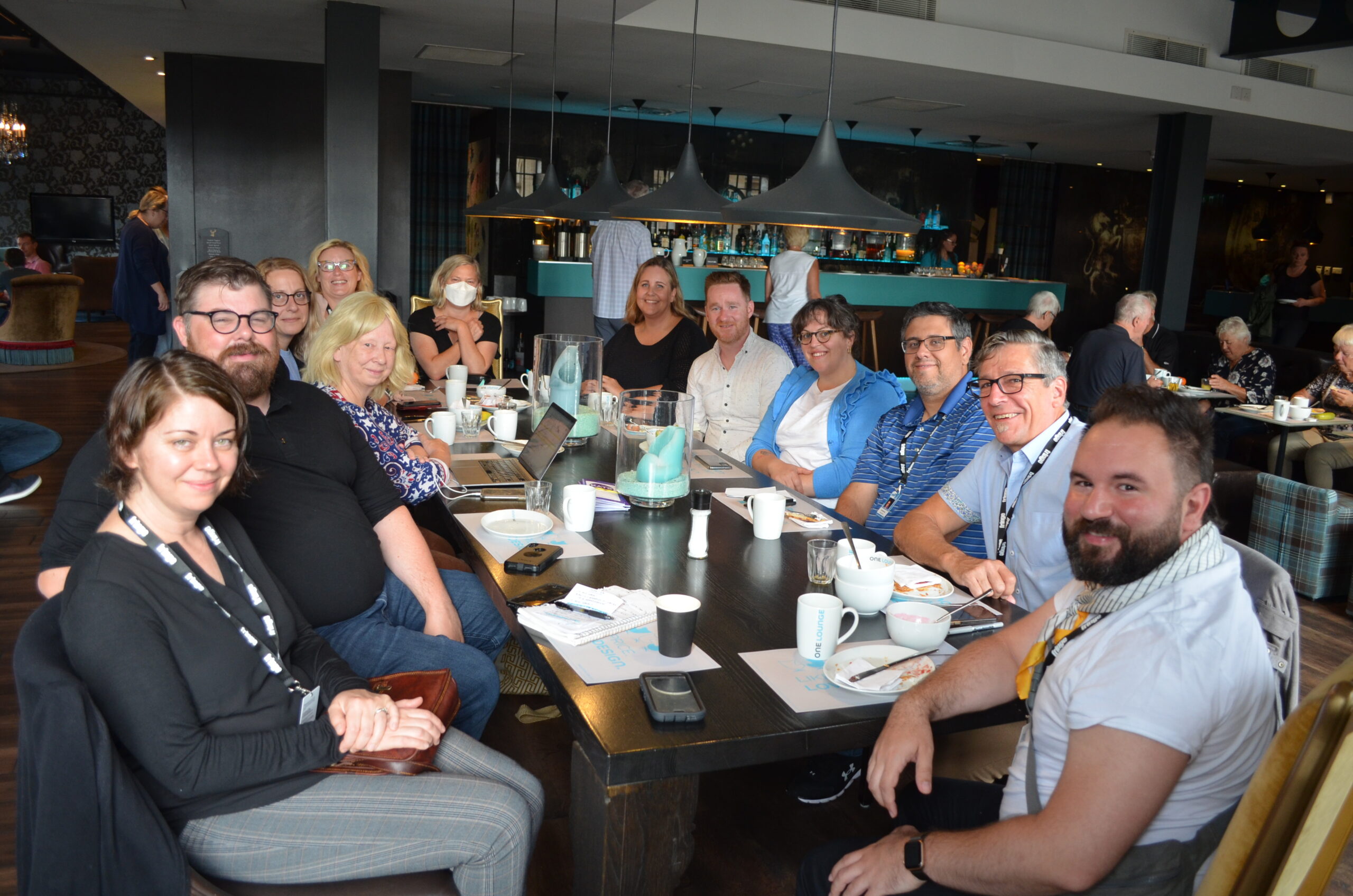 Dans un restaurant bondé, un groupe de délégués est assis autour d'une grande table en souriant pour une photo. Le(la) photographe prend la photo du bout de la table.