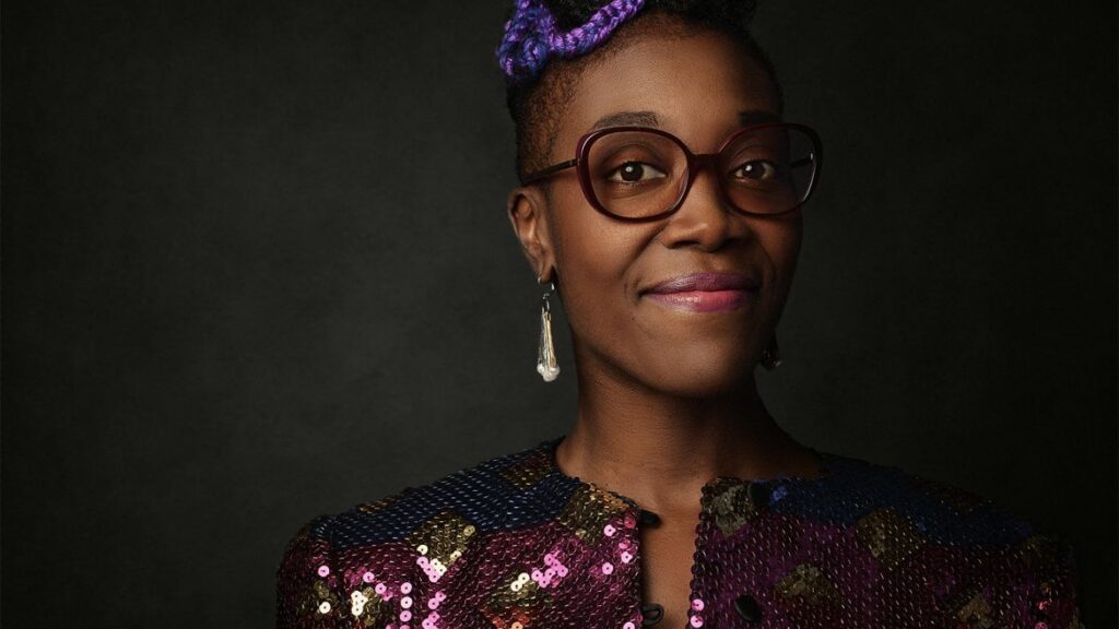 Rhodnie is standing in front of a dark backdrop, smiling towards the camera.