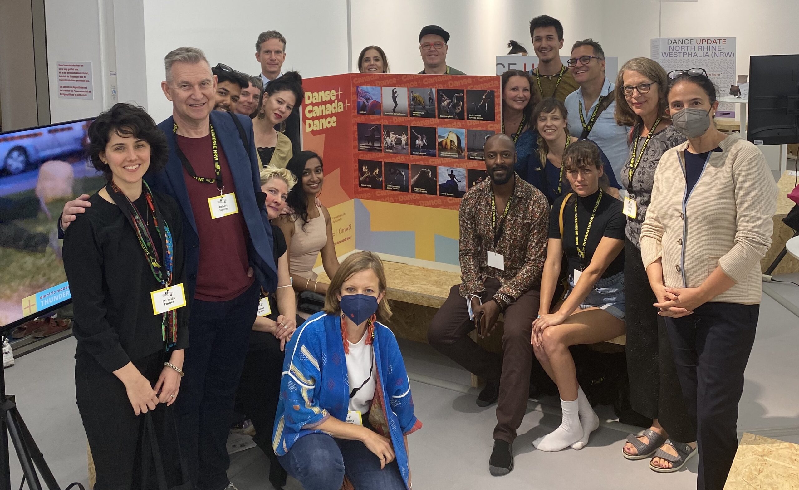 Members of the Canadian delegation at tanzmesse 2022. Some folks are standing, some sitting, and some kneeling around the "Danse + Canada + Dance" booth in the Agora.