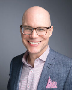 White man in a suit and wearing glasses.