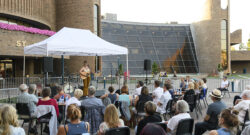 A group of audience members sitting outside in front of the Arden Theatre, watching a musician performing under a tent.
