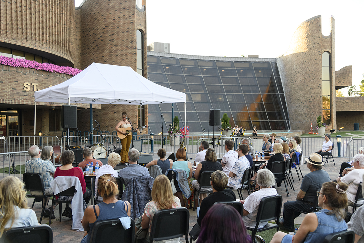 A group of audience members sitting outside in front of the Arden Theatre, watching a musician performing under a tent.