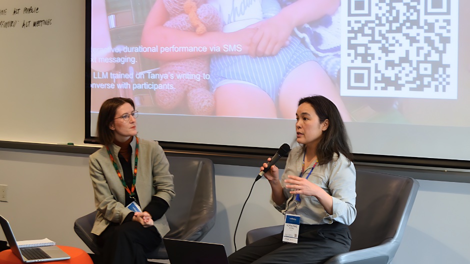 Two women sit in front of a projector screen with laptops in front of them. One woman listens intently while the other speaks into a microphone.