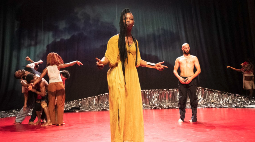 Dobet Gnahoré stands centre stage with arms open wide, with fellow performers standing and performing behind, posing C la Vie’s central question: “How to be together?”