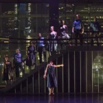 A group of dancers standing on a staircase on a dark stage.