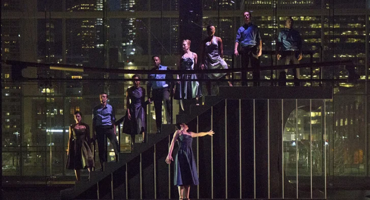 A group of dancers standing on a staircase on a dark stage.