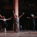 Woman singing with both arms raised in the air, surrounded by four dancers.