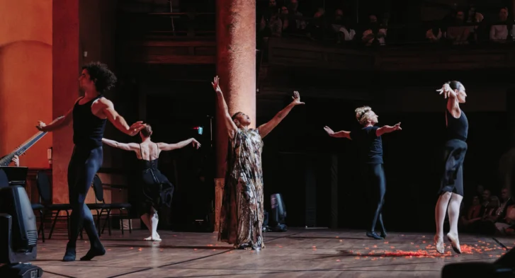 Woman singing with both arms raised in the air, surrounded by four dancers.