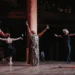 Woman singing with both arms raised in the air, surrounded by four dancers.