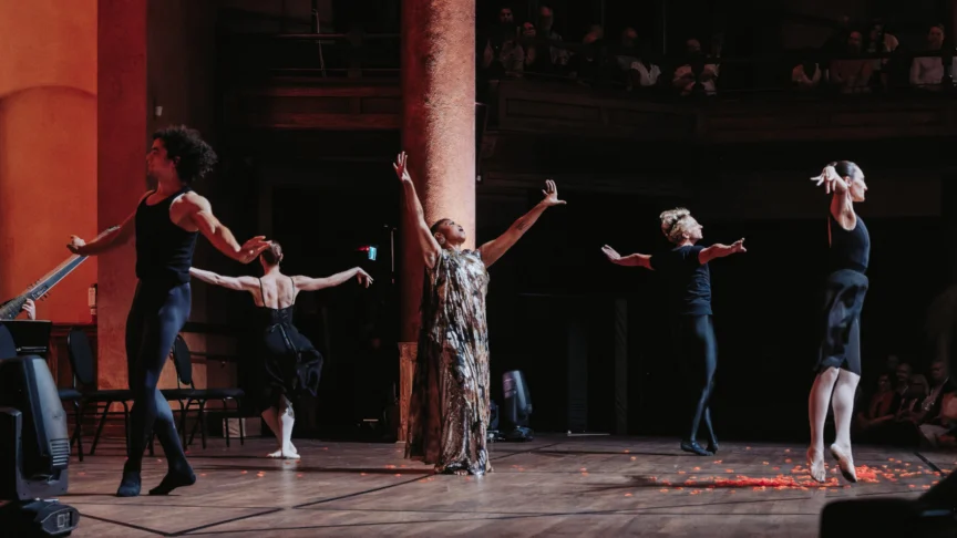 Woman singing with both arms raised in the air, surrounded by four dancers.
